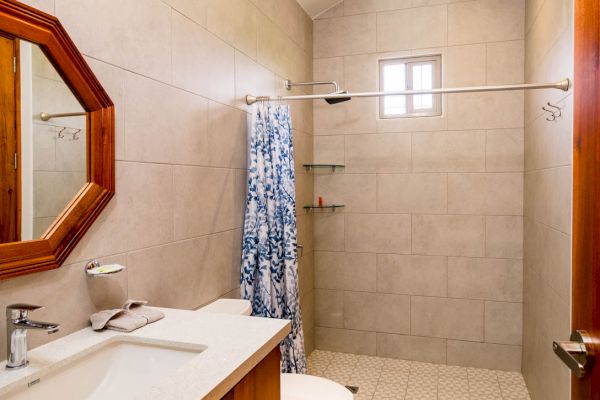 The image shows a modern bathroom with a wooden vanity, white sink, toilet, and shower area with a patterned curtain, under a high ceiling.