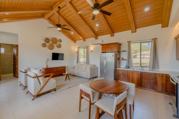 A modern living and dining area with wooden ceiling, two ceiling fans, a TV, sofas, kitchen appliances, and a small dining table with four chairs.