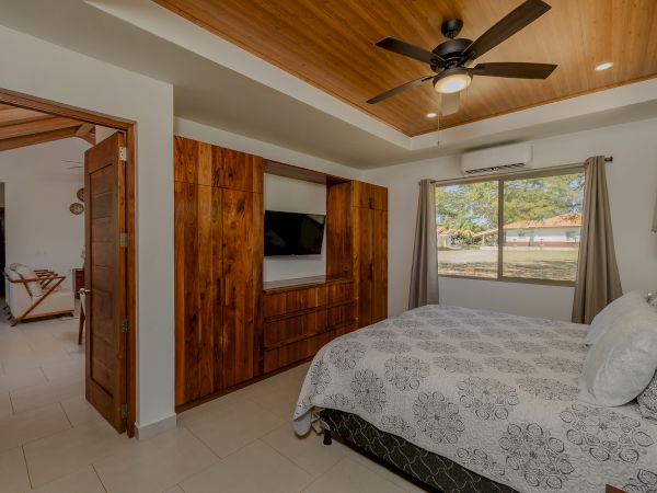 This image shows a cozy bedroom with a patterned gray bedspread, a wooden wardrobe with a TV, a ceiling fan, and a window with a view.