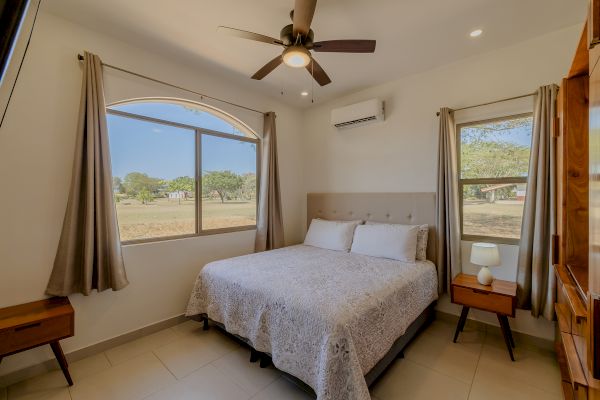 A bedroom with a bed, side tables, lamps, ceiling fan, air conditioner, arched window, curtains, and outdoor view is shown in the image.