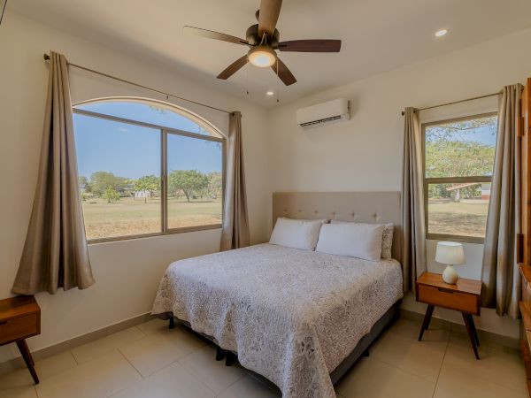 A bedroom with a bed, side tables, lamps, ceiling fan, air conditioner, arched window, curtains, and outdoor view is shown in the image.