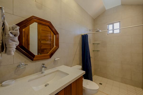 This image shows a modern bathroom with a sink, an octagonal mirror, a toilet, a shower area with a small window, and beige tiles throughout the space.