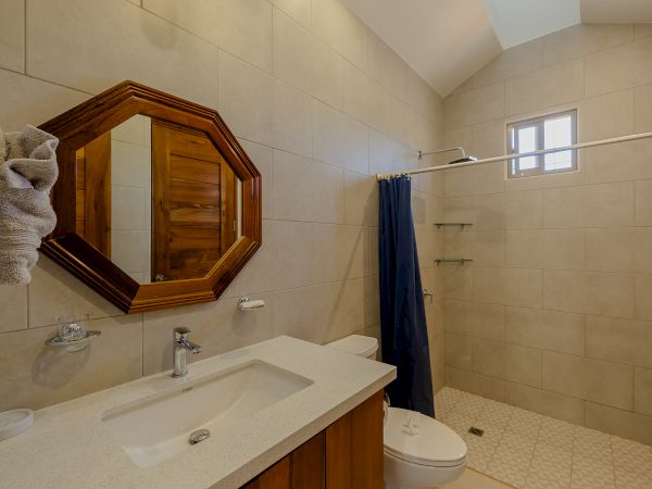 This image shows a modern bathroom with a sink, an octagonal mirror, a toilet, a shower area with a small window, and beige tiles throughout the space.