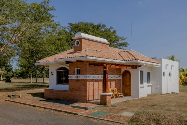 A small, charming brick and white house with a tiled roof and a wooden front door stands in a grassy yard with trees in the background.