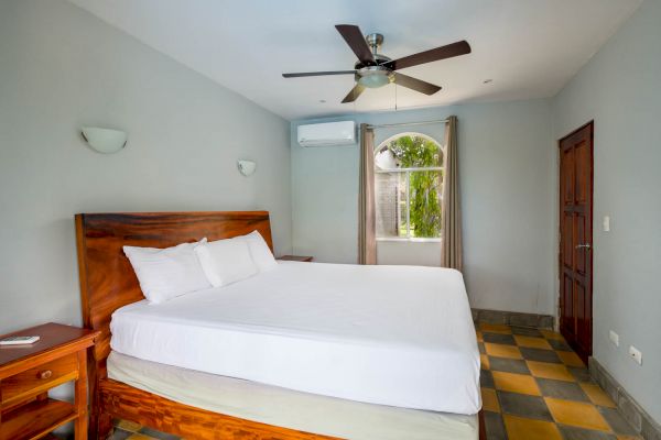 A bedroom with a wooden bed frame, white bedding, a ceiling fan, a window, and a door, all in a simple, clean design.