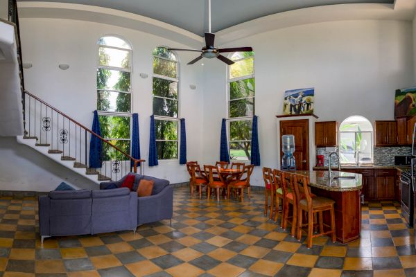 A modern living room and kitchen with checkerboard flooring, a gray couch, wooden dining table, barstools, large windows, and a staircase.