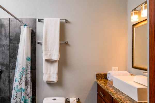 A bathroom with a toilet, sink, mirror, towel rack with towels, shower with curtain, and vanity with granite countertop.