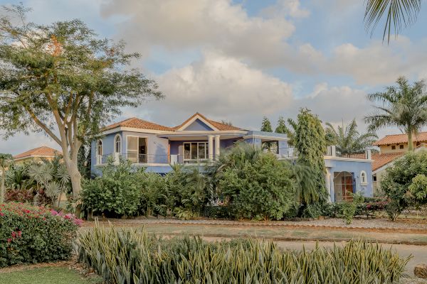 A two-story blue house surrounded by lush greenery, trees, and shrubs under a partly cloudy sky.