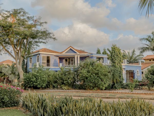 A two-story blue house surrounded by lush greenery, trees, and shrubs under a partly cloudy sky.