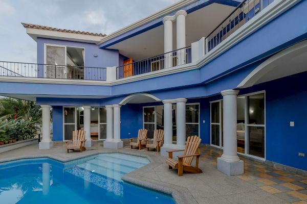 A modern two-story house with blue walls, white columns, balcony, multiple windows, patio seating, and a swimming pool in the foreground.