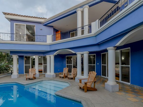 A modern two-story house with blue walls, white columns, balcony, multiple windows, patio seating, and a swimming pool in the foreground.