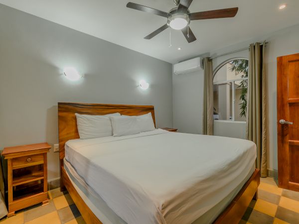 A bedroom with a large bed, two bedside tables, a ceiling fan, wall lights, a wooden door, curtains, and an air conditioning unit on the wall.
