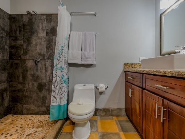 A bathroom with a tiled shower, white toilet, wooden vanity with granite countertop, sink, towel rack, and large mirror.