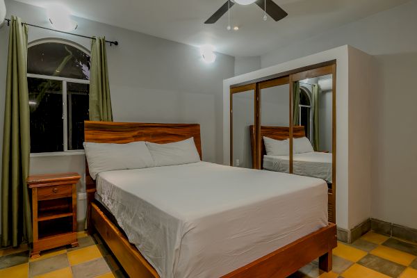 The image shows a bedroom with a bed, wooden headboard, bedside table, large mirrored wardrobe, green curtains, and ceiling fan. There are checkered floors.