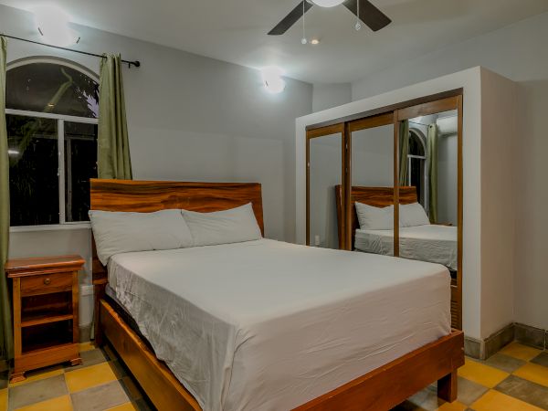 The image shows a bedroom with a bed, wooden headboard, bedside table, large mirrored wardrobe, green curtains, and ceiling fan. There are checkered floors.