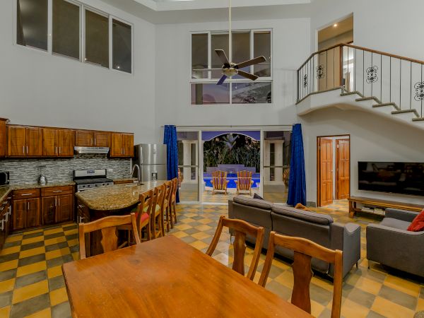 A spacious living area with a kitchen, dining table, sofa, TV, staircase, and balcony, featuring wooden furniture and checkered flooring.