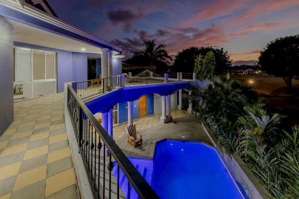 The image shows a modern house with a balcony overlooking a lit swimming pool at dusk. There are palm trees and a scenic sunset in the background.