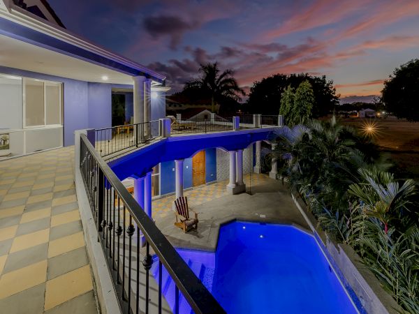 The image shows a modern house with a balcony overlooking a lit swimming pool at dusk. There are palm trees and a scenic sunset in the background.