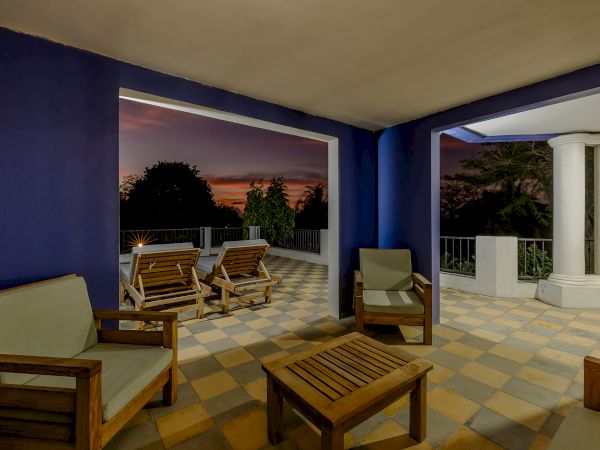 This image shows a patio with wooden furniture and cushioned seating, set against a blue wall, leading to an open view at sunset.