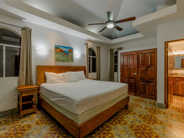 A bedroom with a large bed, ceiling fan, wooden furniture, patterned floor tiles, and an open door leading to a bathroom is shown.