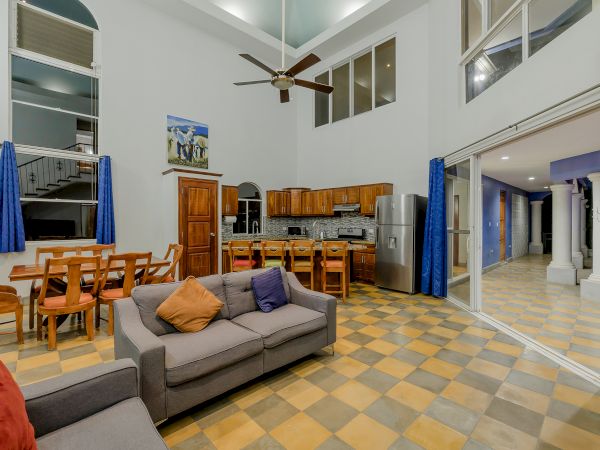 A spacious living area with high ceilings, a ceiling fan, a grey sofa, a dining table with chairs, and a kitchen with wooden cabinets and a stainless steel fridge.