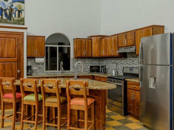 A modern kitchen with wooden cabinets, a stainless steel fridge, an oven, a microwave, an island with four chairs, and colorful cushions.
