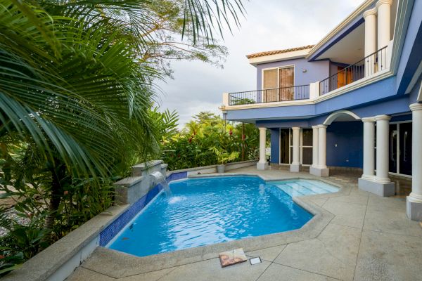 A modern house with a balcony, featuring a blue-painted exterior and a swimming pool surrounded by tropical plants and columns, under a cloudy sky.