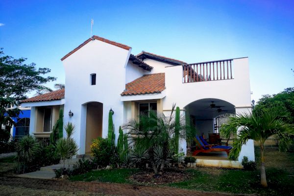 This image shows a white two-story house with a terracotta-tiled roof, surrounded by greenery and small trees, featuring a porch and an upper terrace.