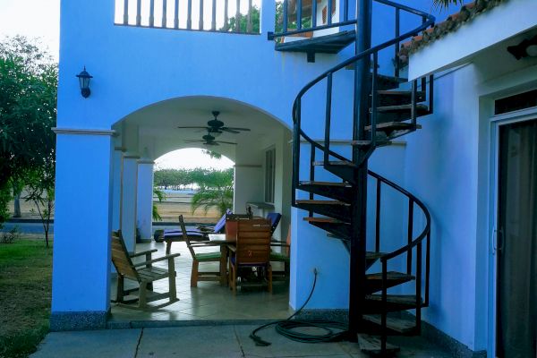 The image shows a two-story building with a spiral staircase, a covered patio with a table and chairs, and an outdoor seating area under a tree.