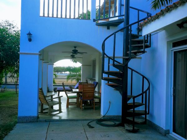 The image shows a two-story building with a spiral staircase, a covered patio with a table and chairs, and an outdoor seating area under a tree.