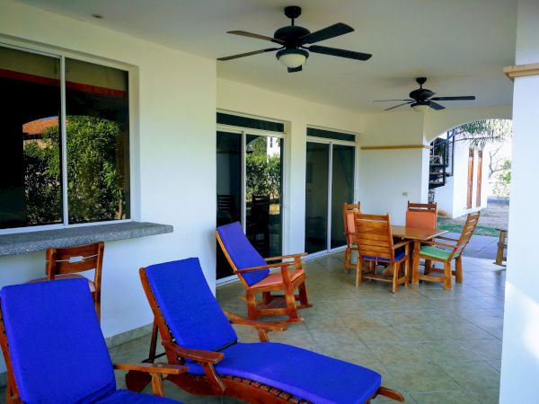 The image shows a patio with blue-cushioned lounge chairs, a wooden dining set, ceiling fans, and large sliding glass doors, suggesting an outdoor relaxation area.