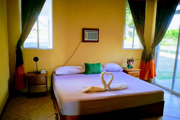 The image shows a cozy bedroom with yellow walls, a double bed, two side tables, an air conditioner, and a folded towel sculpture on the bed.