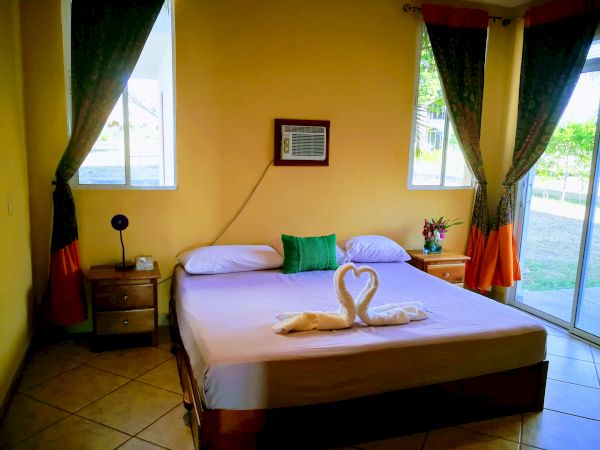 The image shows a cozy bedroom with yellow walls, a double bed, two side tables, an air conditioner, and a folded towel sculpture on the bed.