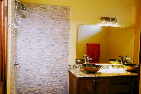 This image shows a bathroom with a tiled shower area, a large mirror above a wooden vanity with a sink, and soft yellow walls.