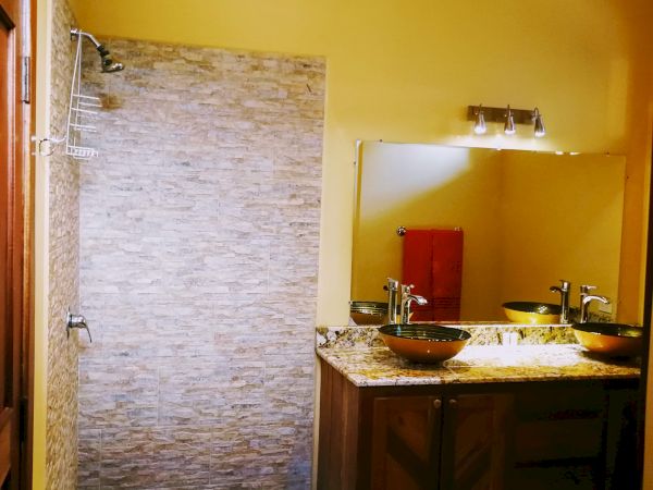 This image shows a bathroom with a tiled shower area, a large mirror above a wooden vanity with a sink, and soft yellow walls.