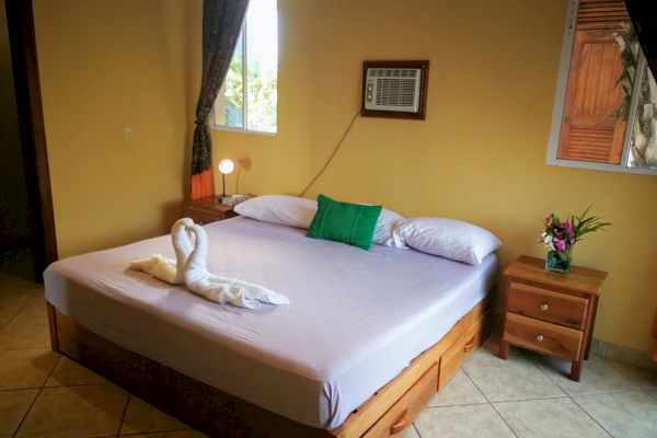 A neatly made bed with swan-shaped towels, a green pillow, a bedside table with a lamp, flowers, and a wall-mounted air conditioner are shown.