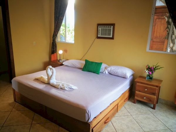 A neatly made bed with swan-shaped towels, a green pillow, a bedside table with a lamp, flowers, and a wall-mounted air conditioner are shown.