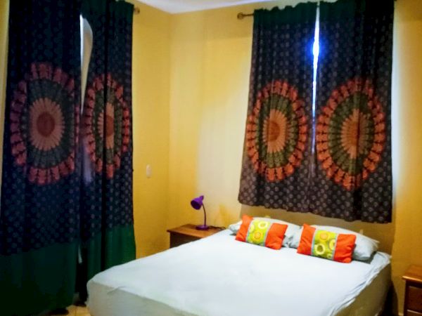A bedroom with a bed, colorful pillows, nightstands, dark curtains with circular patterns, and a ceiling fan. The walls are light yellow.