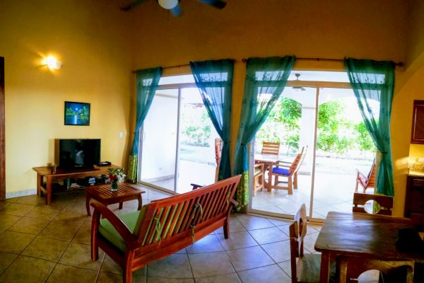 A cozy living room with a TV, wooden furniture, and green curtains, opening up to a patio with outdoor seating and a garden view through sliding doors.