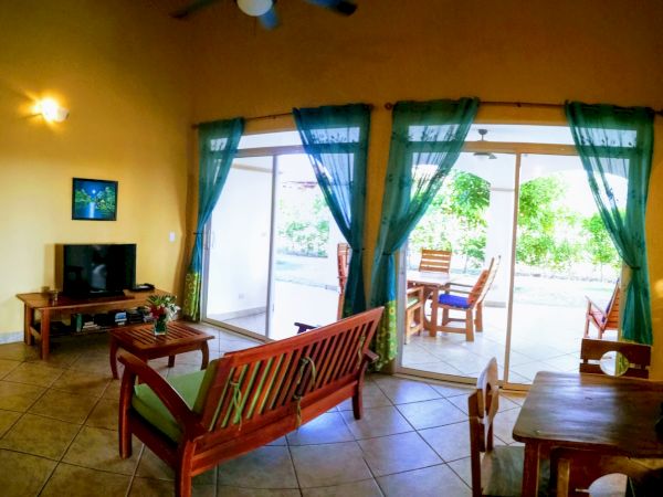 A cozy living room with a TV, wooden furniture, and green curtains, opening up to a patio with outdoor seating and a garden view through sliding doors.