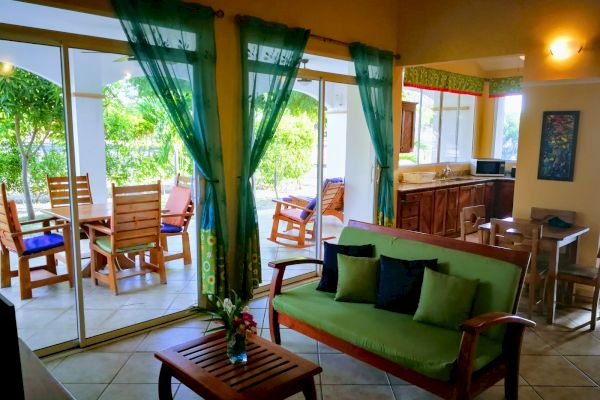 A cozy living room with a green couch, adjacent kitchen, and a patio with wooden furniture, viewed through large glass doors with green curtains.