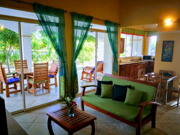 A cozy living room with a green couch, adjacent kitchen, and a patio with wooden furniture, viewed through large glass doors with green curtains.