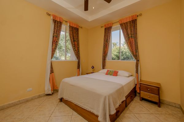 A bedroom with a double bed, two windows with patterned curtains, two bedside tables, and a beige-tiled floor.