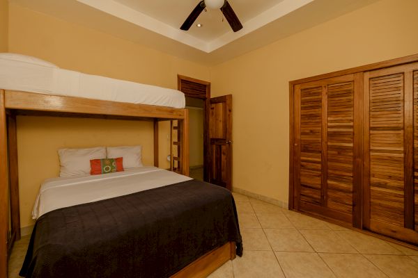 A bedroom with a wooden bunk bed featuring a double bed below and a single bed above, a wooden closet, a wooden door, and a ceiling fan.