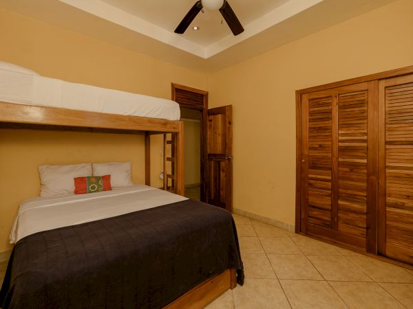 A bedroom with a wooden bunk bed featuring a double bed below and a single bed above, a wooden closet, a wooden door, and a ceiling fan.