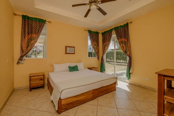 A cozy bedroom features a large bed with green and white pillows, wooden nightstands, decorative curtains, a ceiling fan, and a window view.