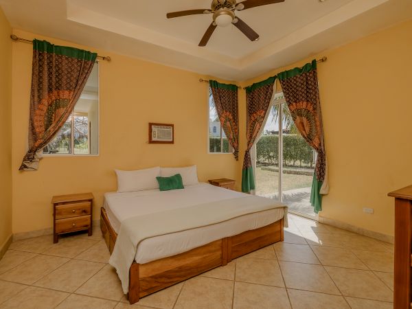 A cozy bedroom features a large bed with green and white pillows, wooden nightstands, decorative curtains, a ceiling fan, and a window view.