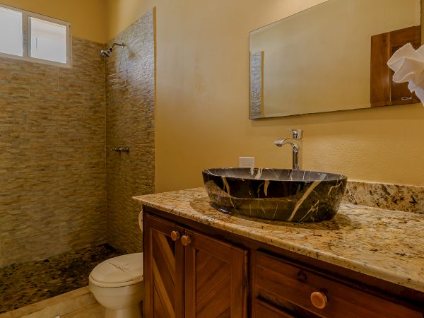 The image shows a bathroom with a wooden vanity, granite countertop, vessel sink, mirror, toilet, walk-in shower, and tile flooring.
