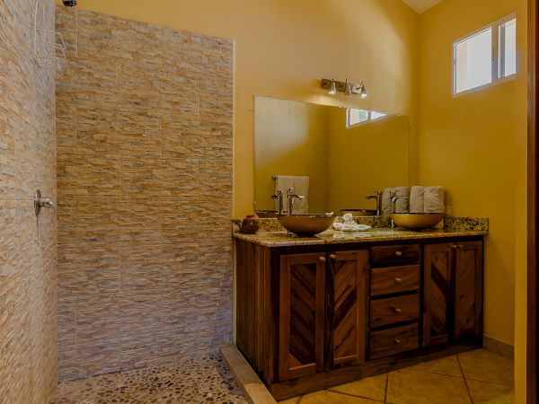 The image shows a bathroom with a walk-in shower, natural stone tiles, a wooden vanity with two sinks, and a large mirror above the vanity.
