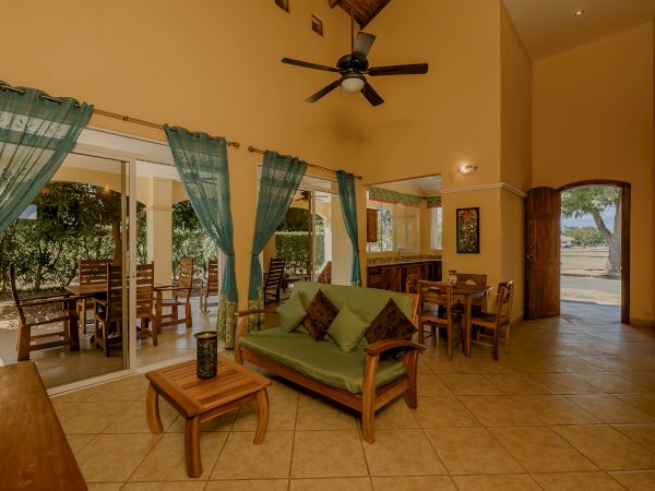 A cozy living area with tall ceilings, a green sofa, wooden furniture, teal curtains, and a view of an outdoor seating patio.